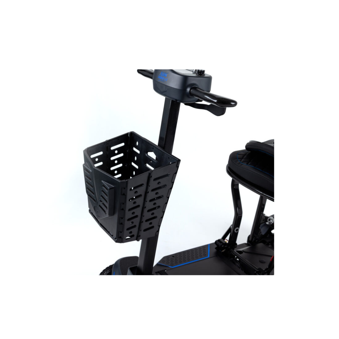 Close-up of a black, perforated Quick Release Folding Basket For Monarch Genie Scooters attached to the mobility scooter's handlebar, with part of the seat visible in the background against a white backdrop.